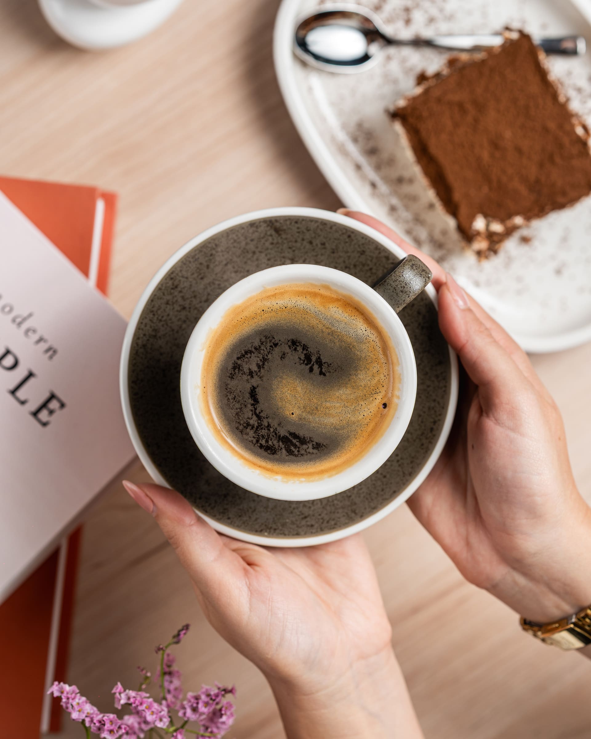 Two hands cradling a stoneware cup of black coffee, a slice of cocoa-dusted tiramisu on the table beside it