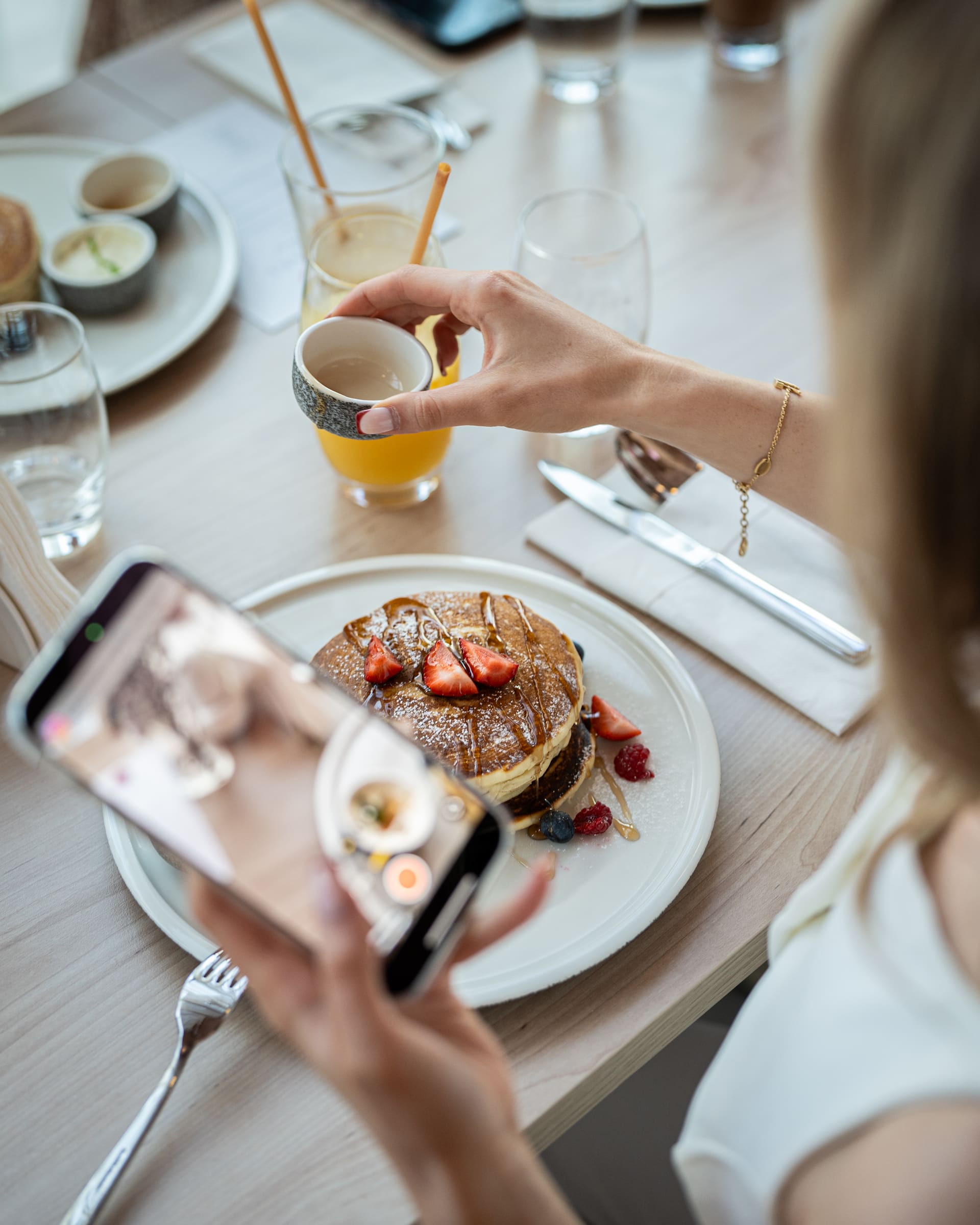 A stack of pancakes with strawberries and berries being photographed on a phone at a sunlit table