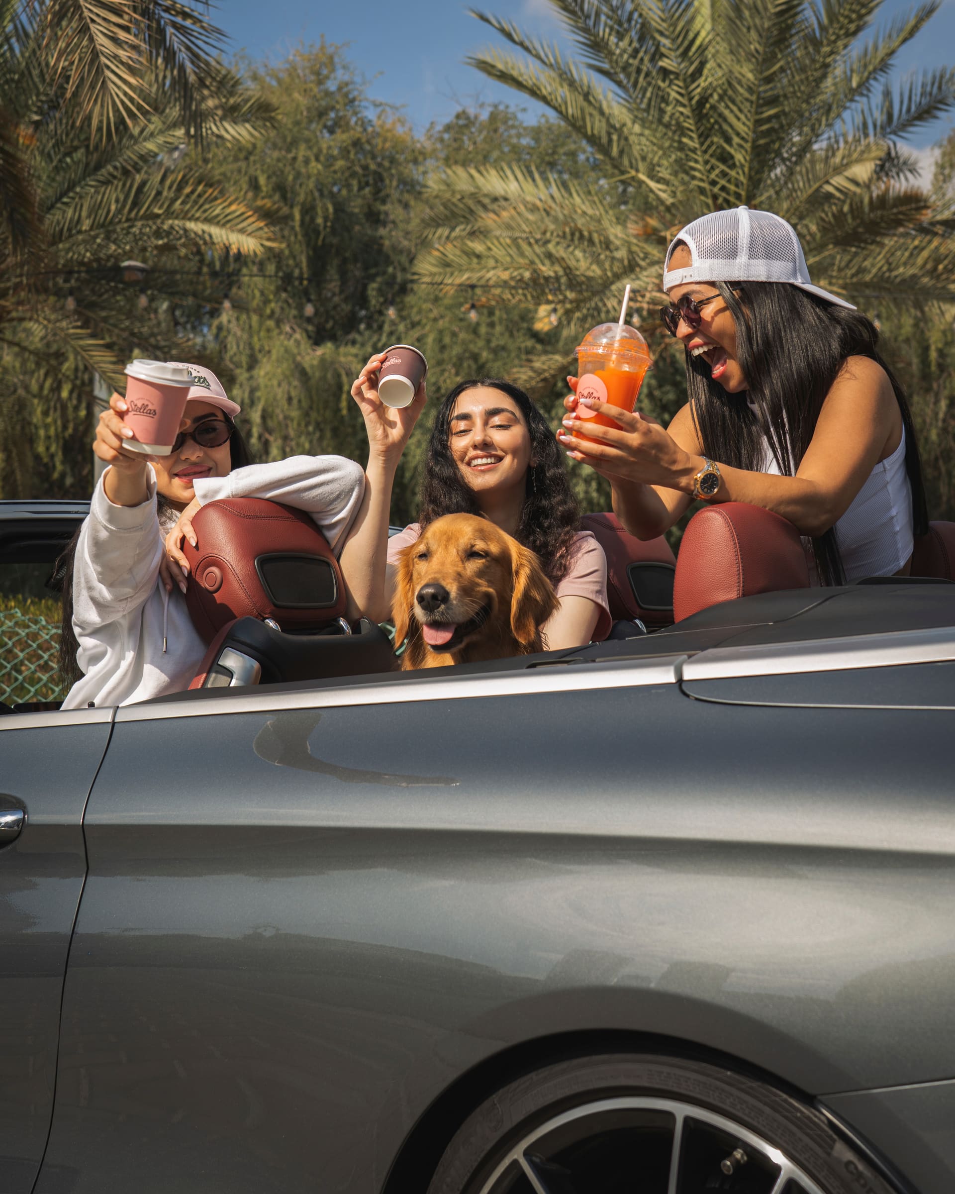 Stella riding in a convertible with three friends holding Stella's Coffee House drinks, palm trees behind them